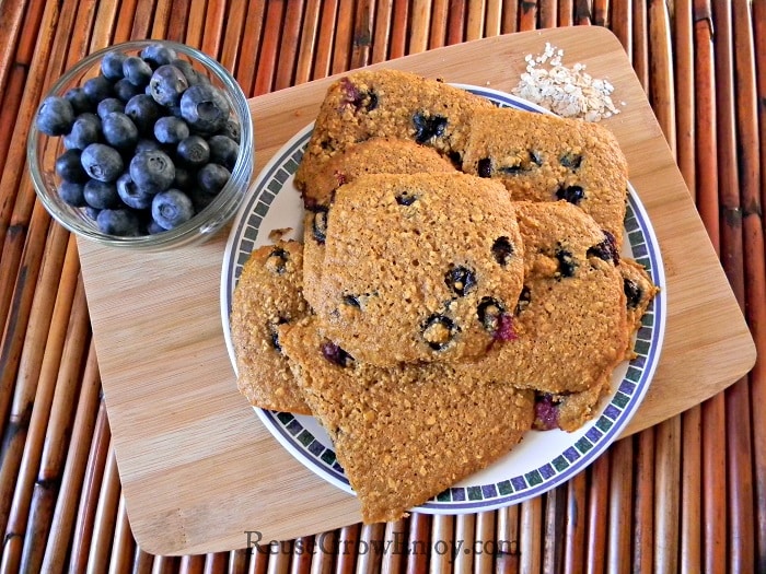 Blueberry oatmeal cookies on white plate with blue trim sitting on a wood cutting board with bowl of fresh blueberries to left side