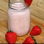 Mason jar filled with strawberry and pineapple protein smoothie with fresh strawberry on side of mouth of jar and some laying beside the jar