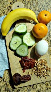 A wood cutting board with egg, jerky, sunflower seeds, dates, banana, cucumber slices and oranges for Paleo snack ideas