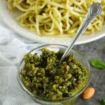 Small glass bowl of homemade pesto with plate of pasta in background.