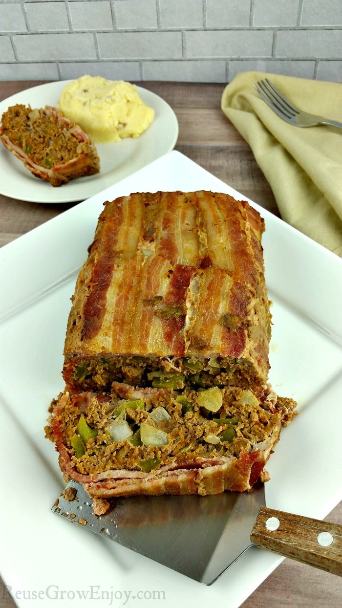 Bacon wrapped Whole30 meatloaf on a white plate. In background is a slice of meatloaf on a small white plate with a serving of mashed potatoes.