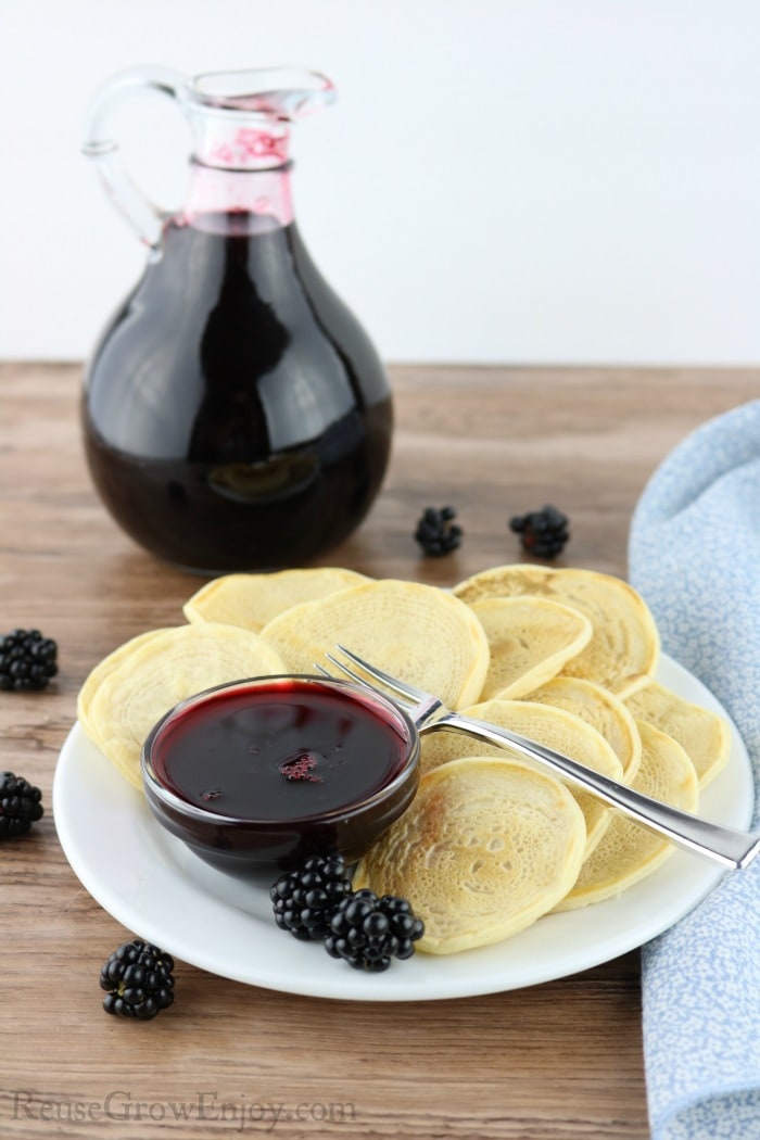 Bottle of blackberry syrup in background. Front is a white plate with bite size pancakes and a dish of the syrup