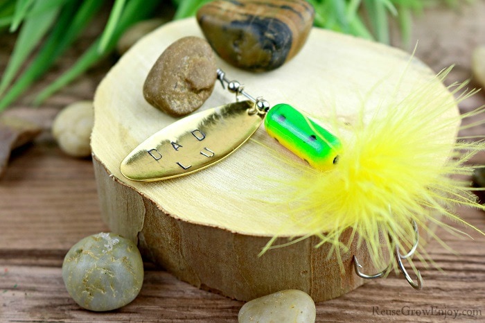 Hand stamped fishing lure laying on wood with rocks.