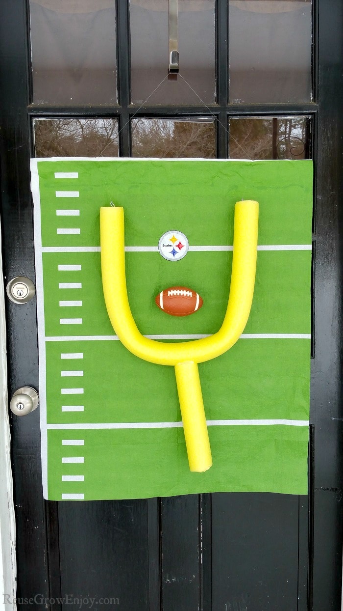 A black door with glass windows with a homemade football decor hanging on it. The decor is a yellow pool noodle making up the goal and a green backing that looks like a football field with a Steelers logo in the center.