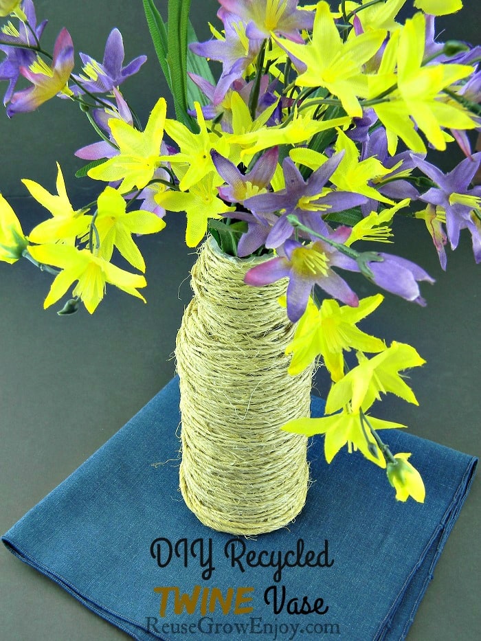 Plastic bottle wrapped in twine and fake flowers in bottle. Setting on a blue cloth. Text overlay at the bottom that says "DIY Recycled Twine Vase"