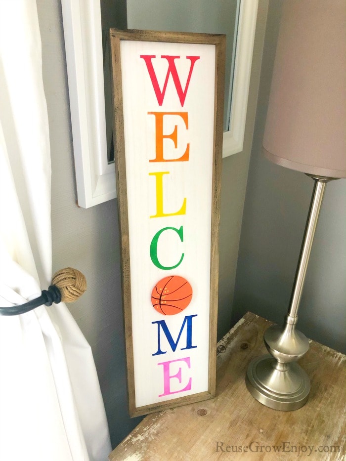 DIY welcome sign displayed next to a window on a wood table with a lamp