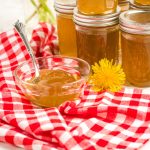 Jars of Dandelion Jelly stacked in background small glass dish of it on a red checkered cloth in the front