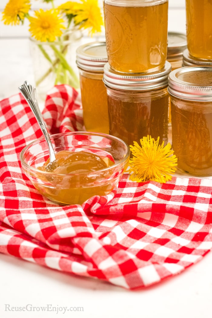 Jars of Dandelion Jelly stacked in background small glass dish of it on a red checkered cloth in the front