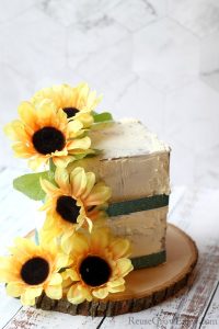 A finished fake cake prop with sunflowers and green ribbon. It is on a wood slice.