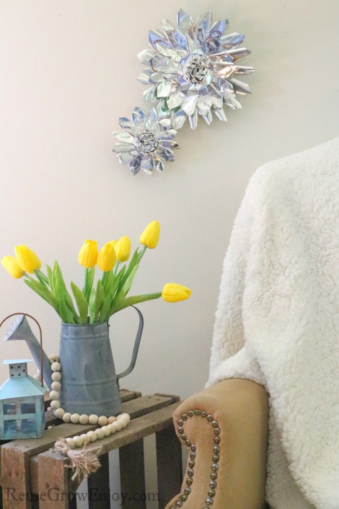 Finished chip bag flowers hanging on wall. Yellow flowers in old watering can on wood table at bottom with farmhouse wood beads.