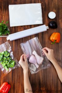 Hands packaging chicken legs in vacuum sealer bag with sealer and produce in background