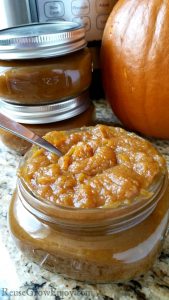 Glass jar full of Instant Pot pumpkin butter with a spoon in it, glass jars, pumpkin shell and Instant Pot in background.