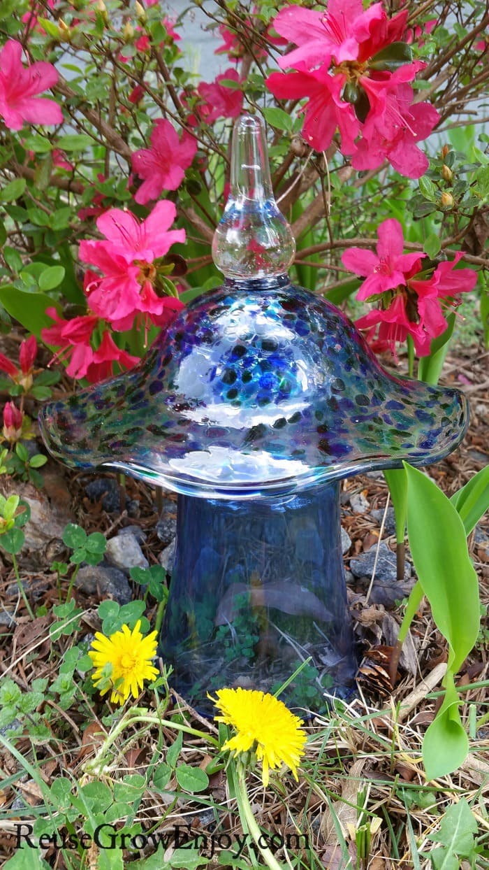 Blue with blue flecks glass garden ornament mushroom made from upcycled glass and candy dish top sitting in front of pink flowers.