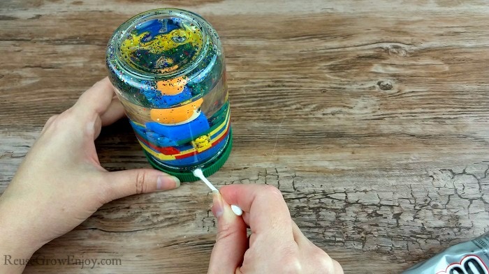 Hand holding q-tip with a dab of glue on it gluing the top to the jar.