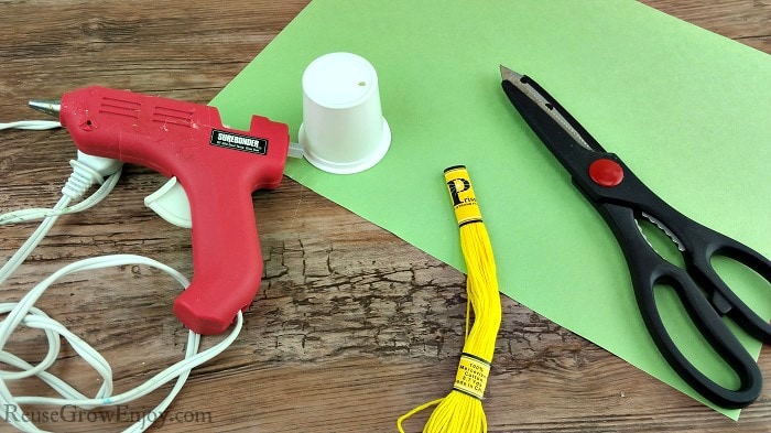 Hot glue gun, yellow string, empty clean k-cup, scissors and green paper.