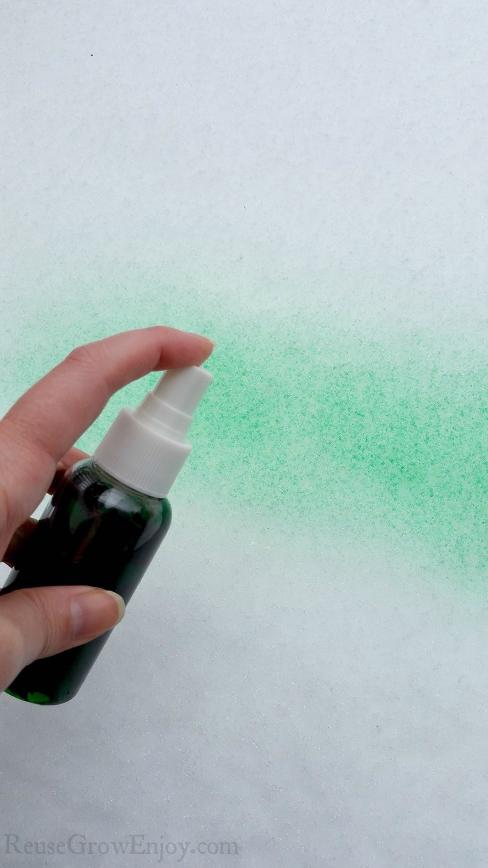 Hand holding small spray bottle with green snow paint being sprayed on snow.