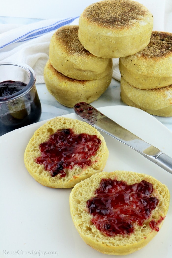open english muffin on white plate with jelly, stack of english muffins and jelly jar in background