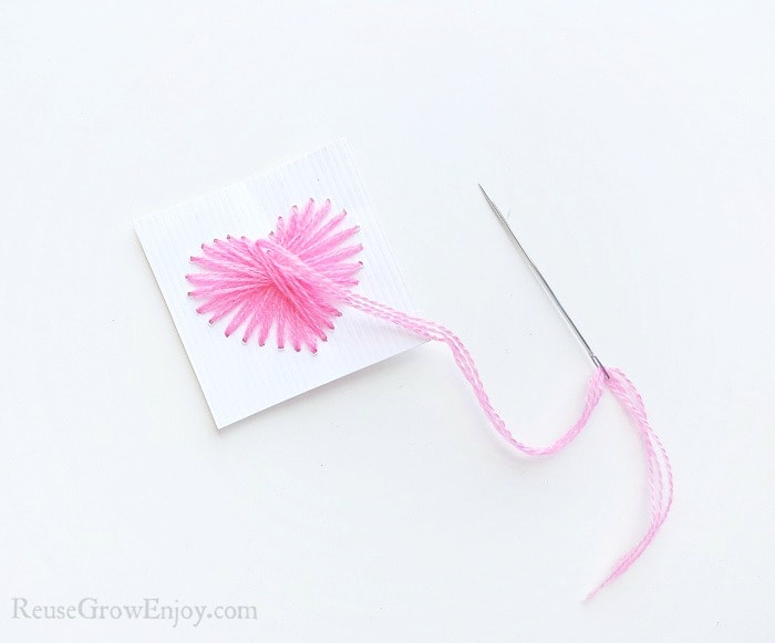 Needle with pink thread on white paper background, small cardstock paper with a heart shape made from holes punched in paper. Pink thread pulled through the holes on the paper.