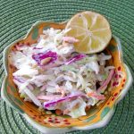 A flower shaped bowl on a light green placemat full of homemade coleslaw with a slice of lemon on the upper right of the bowl.