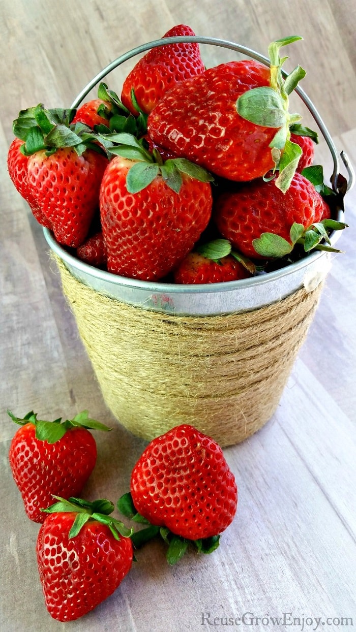 Small tin bucket wrapped in twine filled with fresh strawberries. The fresh strawberries in front of the bucket laying on a wood background.