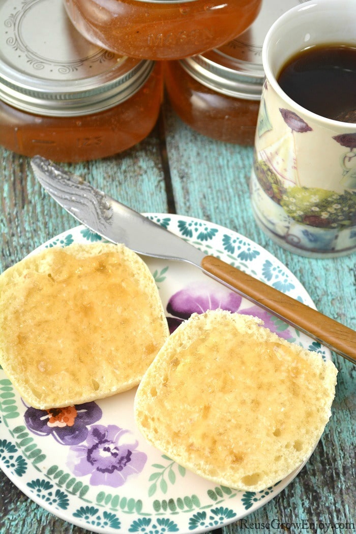 Stacked jars of loquat jelly in the back, cup of coffee to the right side. In the front is biscuit cut in half on a plate with some loquat jelly spread on them with the knife that is laying on the plate.