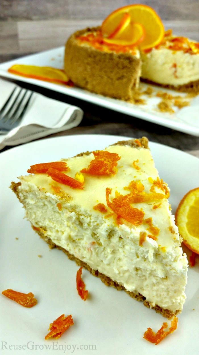 Slice of orange Instant Pot cheesecake in front on a white plate with orange zest as garnish. In the background is the rest of the whole cheesecake.