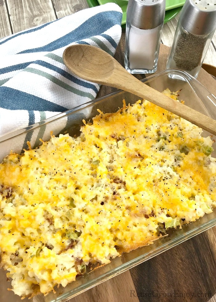 A glass dish full of Keto loaded cauliflower casserole with a wood spoon laying over the top. Blue and white dish towel in back with salt and pepper shakers.