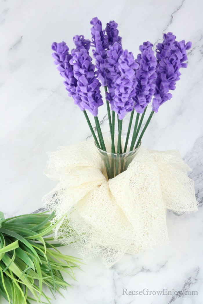 Vase wrapped in lace holding lavender felt flowers with grass bunch to the side