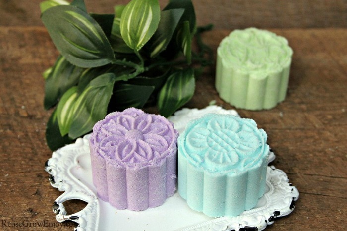 Two press bath bombs on small white tray and one off the tray to the back right. Green leaf plant in background on left.