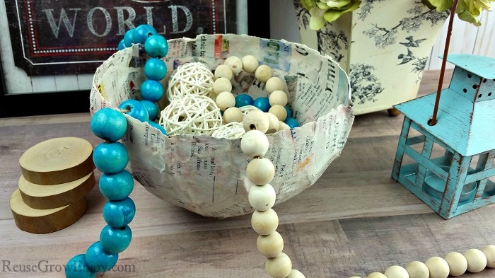 Finished paper mache bowl with farm beads and wicker balls inside. Picture and flowers in background and small teal tealight on the right side.