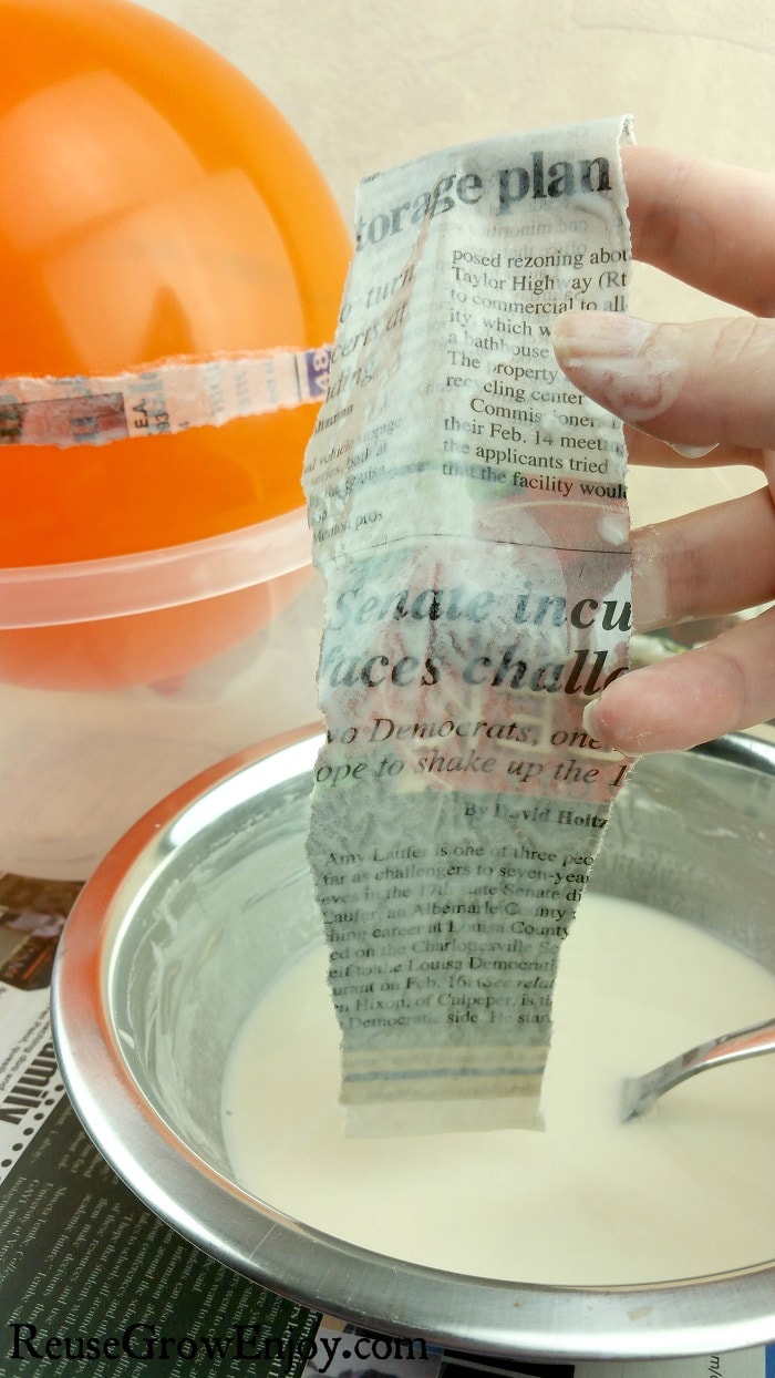 Hand dipping strips of newspaper in paper mache paste with partly covered balloon in background.