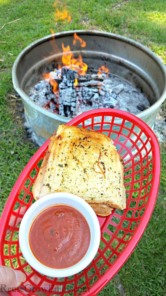 Pepperoni grilled cheese in red basket with a dish or red dipping sauce. Campfire in background.