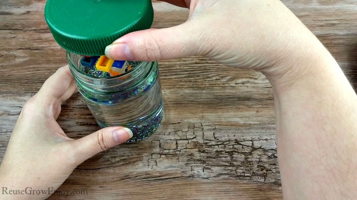 Hand placing top with glued on LEGO's into oil filled jar.