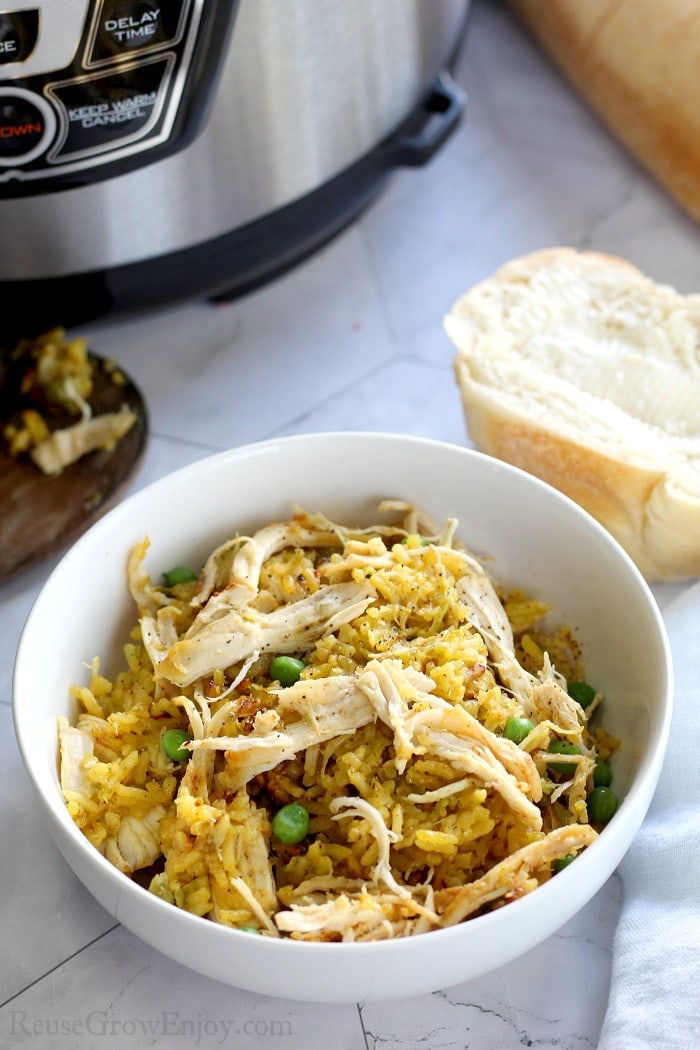 Cooked chicken and yellow rice in a white bowl with a piece of butter bread to the right side and a pressure cooker in the background on the left side.