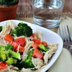 White plate with lemon pepper chicken with broccoli and tomatoes on it with a glass of water in background.