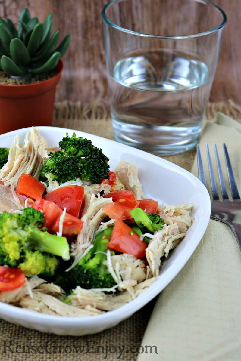 White plate with lemon pepper chicken with broccoli and tomatoes on it with a glass of water in background.