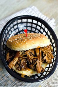 Pulled pork sandwich on a bun sitting in a black plastic basket with a cloth napkin under it and a wood background.