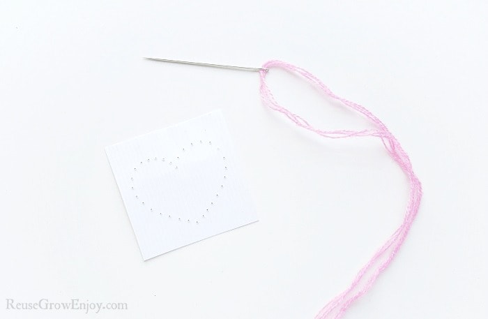 Needle with pink thread on white paper background, small cardstock paper with a heart shape made from holes punched in paper.