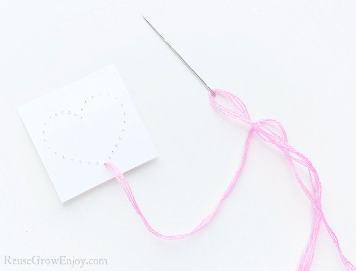 Needle with pink thread on white paper background, small cardstock paper with a heart shape made from holes punched in paper. Pink thread pulled through bottom hole of heart.