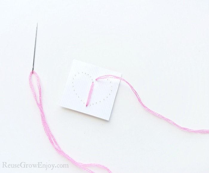 Needle with pink thread on white paper background, small cardstock paper with a heart shape made from holes punched in paper. Pink thread pulled through bottom hole of heart then pulled through the top hole.