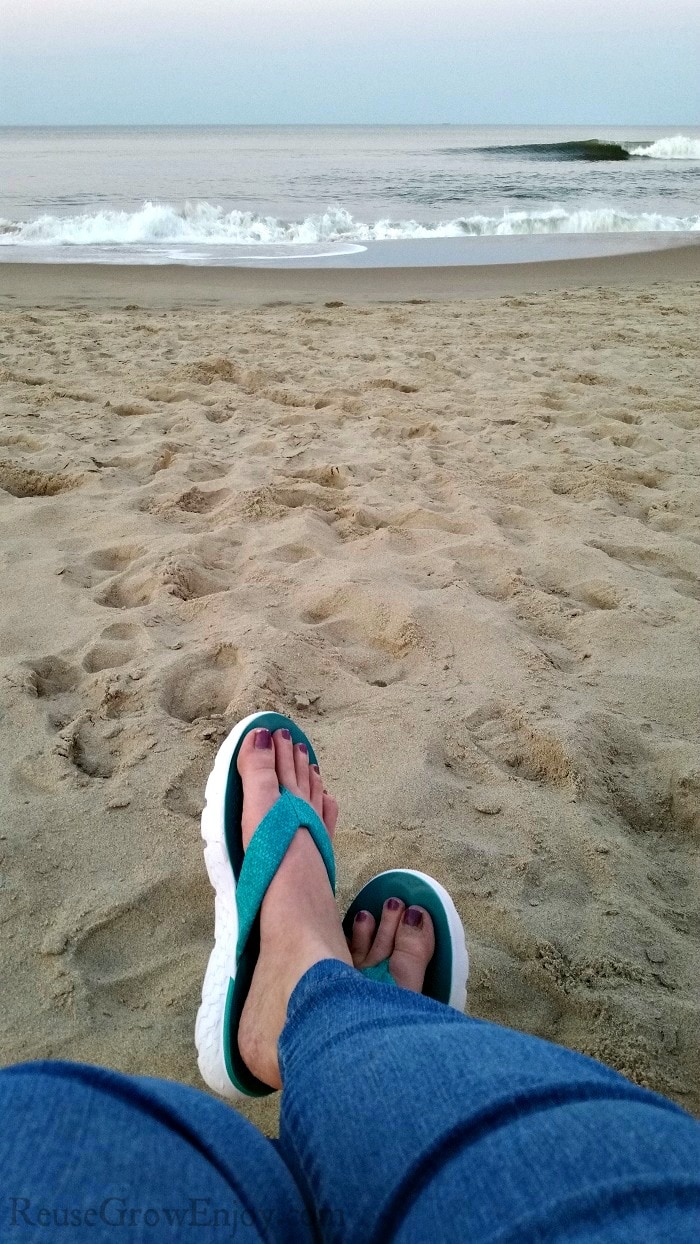 Womens legs in jeans and flip flops crossed with sand and ocean in background.