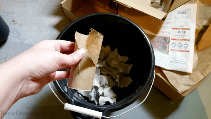 Hand holding a ripped tp tube and black bucket of ripped newspaper in the background.