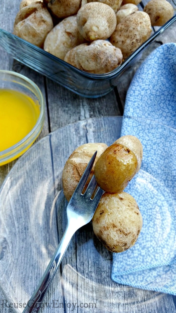 Glass plate with small salt potatoes on it with fork. Dish of butter and bowl of boiled potatoes in background