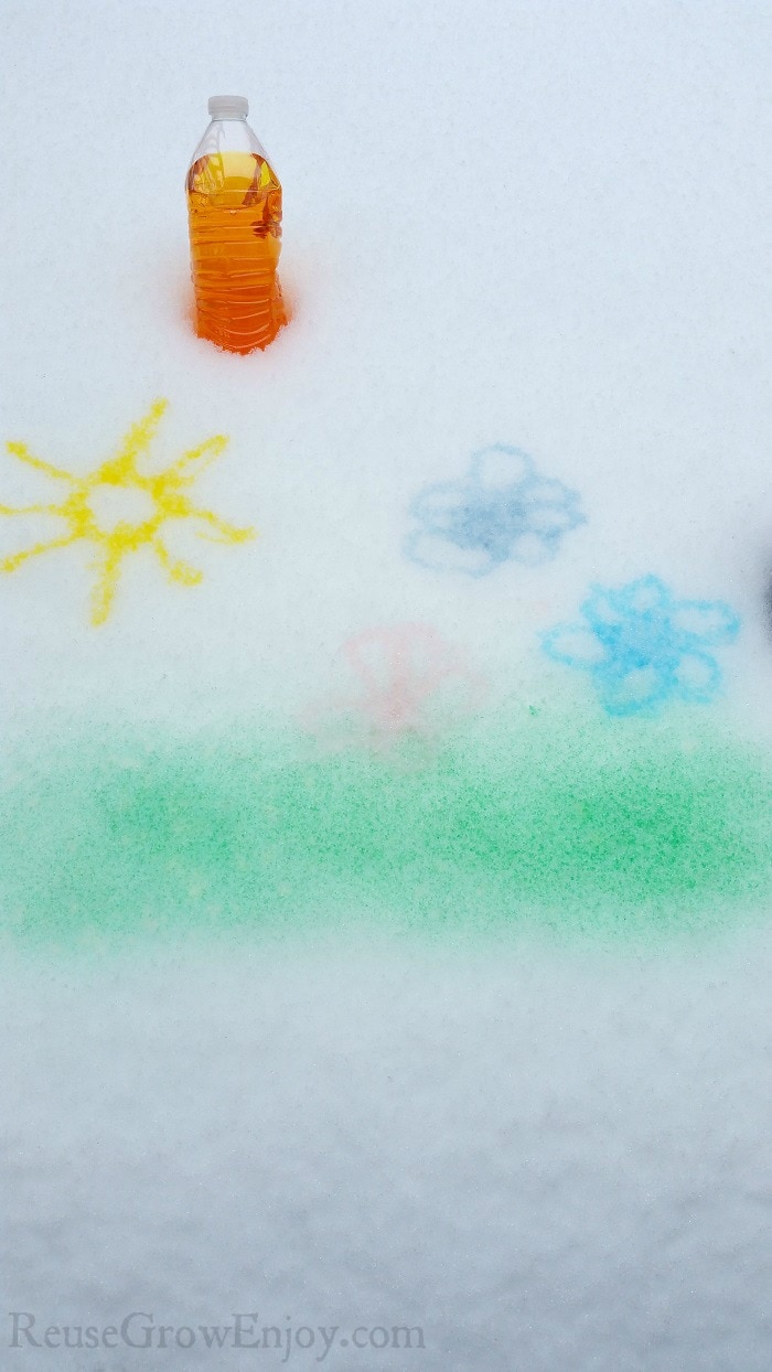 Sun, flowers and grass painted on snow with a bottle of dark yellow snow paint at the top.