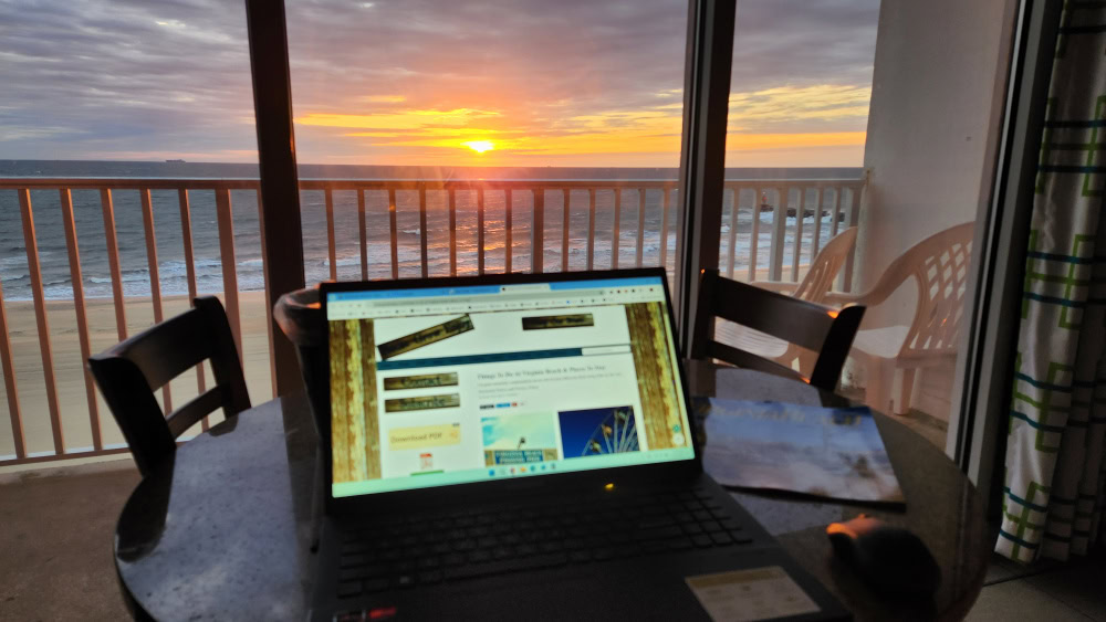 Laptop on table with view of beach sunrise out the window in the background