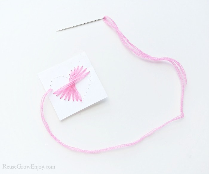 Needle with pink thread on white paper background, small cardstock paper with a heart shape made from holes punched in paper. Pink thread pulled through half the holes on the paper.