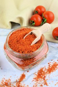 Small glass jar filled with tomato powder with wood scoop on top. Fresh garden tomatoes in background.