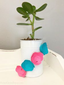 Upcycled can planter that is white with blue and pink paper flowers on it with plant inside. Sitting on a white wood shelf.
