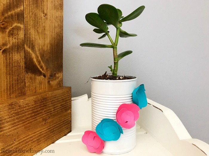 Upcycled can planter that is white with blue and pink paper flowers on it with plant inside. Sitting on a white wood shelf.