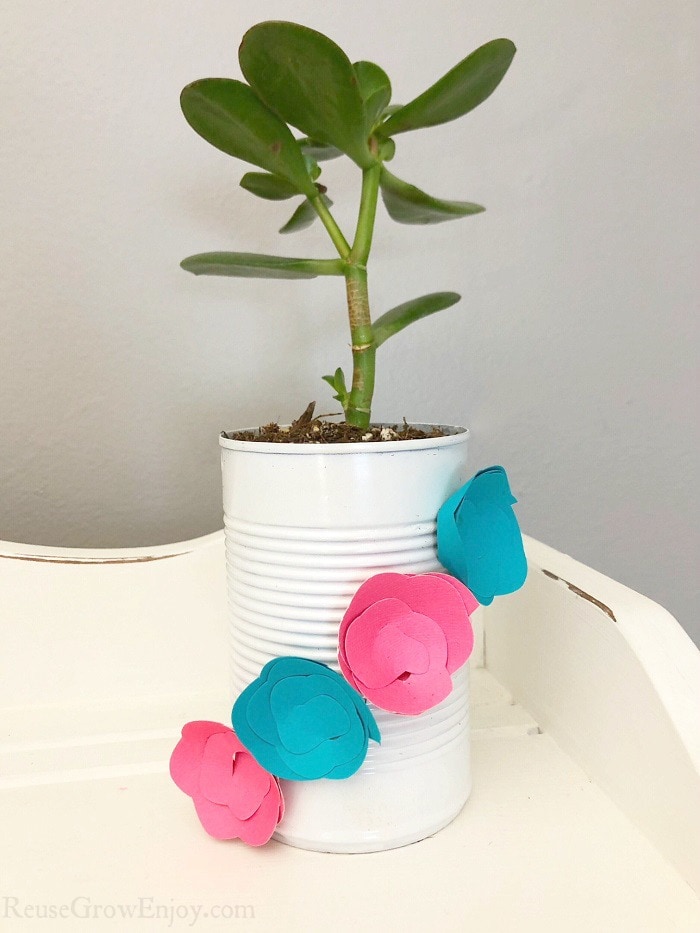 Upcycled can planter that is white with blue and pink paper flowers on it with plant inside. Sitting on a white wood shelf.